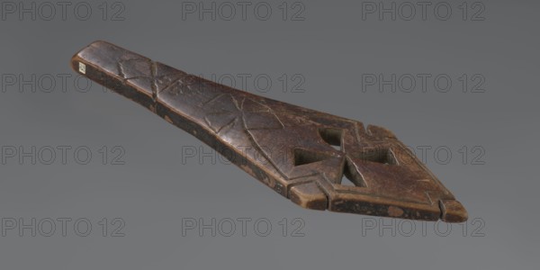 Hand cross fragment, Simple engraved decoration on the surface: four triangular cutouts arranged in the form of a cross; below the holes two rather carelessly engraved crosses; the reverse also decorated with a cross motif. Decoration engraved with a thin, shallow line. Dark wood. 20th century, Ethiopia. Cross; height 26 cm, width 8 cm., A tapered carved wooden object with a pointed end and a flattened wider end, showing a carved geometric pattern that includes cut-out diamond-shaped openings; the surface has worn areas and scratches and displays shades of brown with darker brown and some lighter tan patches, set against a plain gray background.
