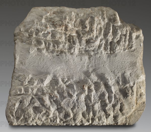 Honorific inscription, unknown workshop of Asia Minor author; Roman legions, military decorations; 120–130; Asia Minor Heraclea Salbake; inscription; height 57 cm, width 49 cm, depth 8 cm, weight 49.5 kg, A roughly rectangular block of stone with an uneven, weathered surface, showing a mix of smoother and heavily textured areas; the stone displays shades of gray and off-white with small patches of brownish rust coloring and subtle darker gray shadows in crevices; the top and lower portions have pronounced ridges and pitted indentations while a central horizontal band appears relatively smoother and slightly lighter; the edges are irregular and worn, and the overall appearance is matte and coarse.