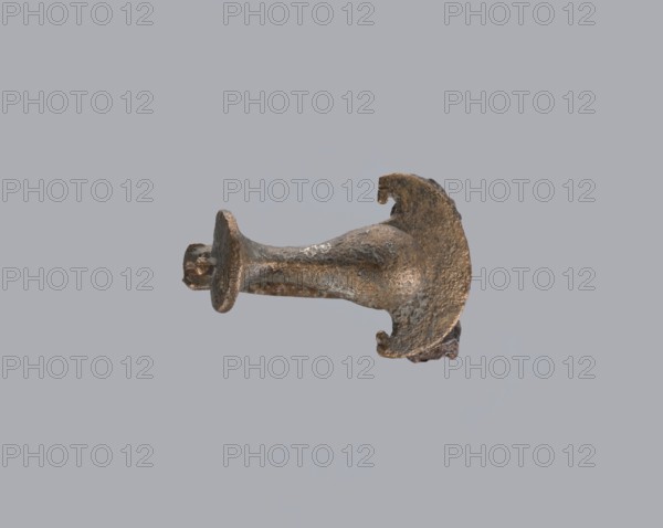 Knuckle-shaped fibula, unknown Roman workshop; fibula with a flat underside, rounded at the top, rising from a horizontal semicircle upwards, bent at an acute angle and terminating in a semicircle with a button in the center. Below the arch, on one side, there is a long spiral from which a pin emerges; on the other, a high stem at the end is bent into a sheath. Material: bronze. Condition: good. Date: 2nd half of the 2nd–1st half of the 3rd century, Roman Empire 27 BC–476. Type: fibula. Dimensions: height 2.6 cm, width 3.7 cm. Provenance note: repossession Silesia., A small metallic object with a rough, corroded surface shown against a plain light gray background; the object appears irregularly shaped with a flared, circular end and a narrower stem leading to a curved, partially broken section, showing textures that include pitting and flaky areas; visible colors are shades of brown, dark bronze, muted gold, and areas of grayish discoloration.