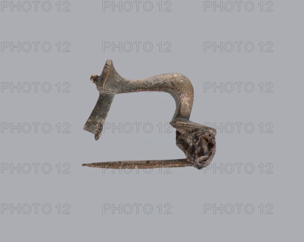 Knuckle-shaped fibula, unknown Roman workshop. Fibula with a flat underside, rounded at the top, rising from a horizontal semicircle upwards, bent at an acute angle and terminating in a semicircle with a button in the center. Below the arch, on one side, there is a long spiral, from which a pin emerges; on the other, a high stem at the end is bent into a sheath. Material: bronze. Condition: good. Date: 2nd half of the 2nd–1st half of the 3rd century. Culture: Roman Empire 27 BC–476. Type: fibula. Dimensions: height 2.6 cm, width 3.7 cm., A single small metal object with a curved upper section and an attached straight pin-like lower piece is displayed against a plain light gray background; the object appears corroded and rough with a patina showing dark brown, dull bronze, and hints of greenish tarnish, and there is a twisted, irregular cluster of metal at one end while the opposite end shows a flattened, broken-looking extension.