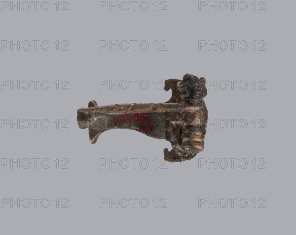 Knuckle-shaped fibula, unknown Roman workshop. Fibula with a flat underside, rounded at the top, rising from a horizontal semicircle upwards, bent at an acute angle and terminating in a semicircle with a button in the center. Below the arch, on one side, there is a long spiral, from which a pin emerges; on the other, a high stem bent into a sheath. Material: bronze. Condition: good. Date: 2nd half of the 2nd–1st half of the 3rd century CE Roman Empire. Type: fibula. Dimensions: height 2.6 cm, width 3.7 cm. Provenance: repossession Silesia., A small metallic object with a weathered, rough surface sits against a plain light gray background, showing a mix of brown, dark bronze, and greenish corrosion tones; one end has a sculpted figure with textured facial features and a hand-like form, while the opposite end extends into a curved, slightly tapered arm with a flattened top and a small rounded protrusion; overall the object appears irregular and aged with areas of darker patina and lighte...