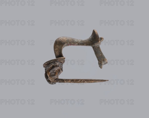 Knuckle-shaped fibula, unknown Roman workshop. Fibula with a flat underside, rounded at the top, rising from a horizontal semicircle upwards, bent at an acute angle and terminating in a semicircle with a button in the center. Below the arch, on one side, there is a long spiral, from which a pin emerges; on the other, a high stem at the end is bent into a sheath. Material: bronze. Condition: good. Date: 2nd half of the 2nd–1st half of the 3rd century, Roman Empire 27 BC–476. Type: fibula. Dimensions: height 2.6 cm, width 3.7 cm., A close-up view of a small metallic object against a plain gray background, showing an L-shaped curved upper piece with a textured, corroded surface in shades of dark brown, rust, and dull bronze, attached to a straight thin rod that extends horizontally and tapers to a pointed end with similar brown and rust tones; the left end of the rod connects to a knotted, irregular cluster of metal with layered, curled folds and rough pitting in dark brown and bronze hue...