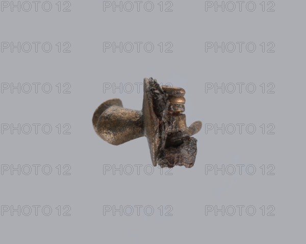 Knuckle-shaped fibula, unknown Roman workshop. Fibula with a flat underside, rounded at the top, rising from a horizontal semicircle upwards, bent at an acute angle and terminating in a semicircle with a button in the center. Below the arch, on one side, there is a long spiral from which a pin emerges; on the other, a high stem at the end is bent into a sheath. Material: bronze. Condition: good. Date: 2nd half of the 2nd–1st half of the 3rd century CE, Roman Empire 27 BC–476. Type: fibula. Dimensions: height 2.6 cm, width 3.7 cm., A small metallic object with a rough, irregular surface floats against a solid pale gray background; the object has a bulbous rounded end and a flatter, jagged section with layered ridges and a curved protrusion, displaying a mix of bronze, dark brown, and black tones with subtle highlights.