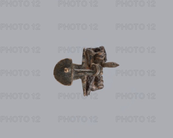 Knuckle-shaped fibula, unknown Roman workshop. Knuckle-shaped fibula with a flat underside, rounded at the top, rising from a horizontal semicircle upwards, bent at an acute angle and terminating in a semicircle with a button in the center. Below the arch, on one side, there is a long spiral from which a pin emerges; on the other, a high stem at the end is bent into a sheath. Condition: good. Date: 2nd half of the 2nd–1st half of the 3rd century CE, Roman Empire 27 BC–476. Material: bronze. Function/type: fibula. Dimensions: height 2.6 cm, width 3.7 cm., A small corroded metal object with a dark brown and greenish patina centered against a pale gray background, showing a rounded head on one end and a pointed tip on the other, a short shaft connecting them, and irregular rough surfaces with flaky texture and spots of lighter brown and faint metallic highlights.