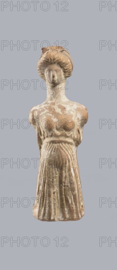 Terracotta figurine of a woman, terracotta, female molded figurine, Greece, orange clay, surface covered with whitewash. Woman dressed in a short, pleated chiton girdled at the waist. Long, thick neck and disproportionately small head with an oval face. Hair combed upwards and tied in a bun, held by a headband tenia tied in a bow at the top of the head. The back is unfinished. Originally had separately attached arms and legs below the knees, indicated by a recess and perforations for hooks. Condition: missing arms and legs, whitewash worn away. Circa 400 BC. Function: toy. Height 10.1 cm, width 3.5 cm., A vertically oriented image of a small worn statue of a standing female figure centered against a plain light gray background; the statue appears made of a weathered brownish-beige material with areas of lighter beige and off-white surface encrustation, showing a rounded head with stylized hair gathered and textured into a high coiffure, a simplified face with smooth, indistinct feature...