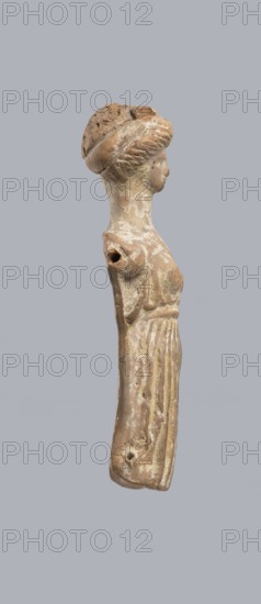 Terracotta figurine of a woman, terracotta, female molded figurine, Greece, orange clay, surface covered with whitewash. Woman dressed in a short, pleated chiton girdled at the waist. Long, thick neck and disproportionately small head with an oval face. Hair combed upwards and tied in a bun, held by a headband tenia tied in a bow at the top of the head. Back of the figurine is unfinished. Originally had separately attached arms and legs below the knees, evidenced by a recess and perforations for hooks. Condition: missing arms and legs, whitewash worn away. c. 400 BC. Toy. Height 10.1 cm, width 3.5 cm., A weathered small sculpted figure in profile against a plain gray background, showing a head with a rounded hairstyle or headpiece and a long slender neck leading to a narrow torso with displaced or damaged arms; the surface has an aged, rough texture with areas of flaking and small holes, and the visible colors are various shades of brown, beige, off-white, and hints of pale yellow.