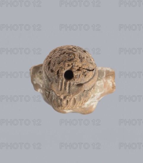 Terracotta figurine of a woman, terracotta, female molded figurine, Greece, orange clay, surface covered with whitewash. Woman dressed in a short, pleated chiton girdled at the waist. Long, thick neck and disproportionately small head with an oval face. Hair combed upwards and tied in a bun, held in place by a headband tenia tied in a bow at the top of the head. The back of the figurine is unfinished. The figurine originally had separately attached arms and legs below the knees, as evidenced by a special recess and perforations for hooks. Condition: missing arms and legs, whitewash worn away. c. 400 BC, Greece, toy. Height 10.1 cm, width 3.5 cm., A weathered brown object with a rough, textured surface and an irregular rounded top that has a circular hole near its center, resting on a lighter brown base with mottled pale patches; the background is a uniform light gray.