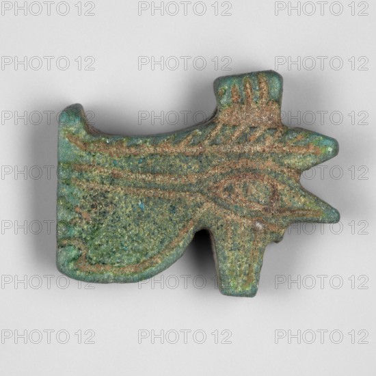 Amulet — udjat eye; maker, udjat eyes; carefully finished amulet in the shape of an udjat eye with a pendant placed above the upper edge of the eyebrow; New Kingdom; found in Edfu; amulet; height 1.3 cm, width 1.7 cm., A small carved object with an irregular shape that suggests an abstract animal head, showing textured surfaces and raised linear grooves; the piece displays a mix of green and turquoise tones with areas of brown and mottled darker green, and the lighting casts a soft shadow beneath it on a pale gray background.