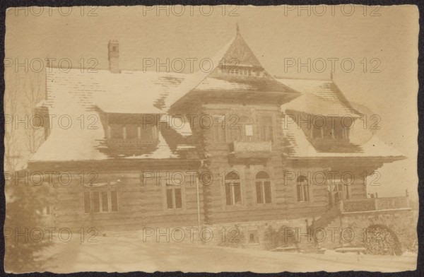 Zakopane. Villa pod Jedlami, built in 1897 for Jan Gwalbert Pawlikowski according to a design by Stanislaw Witkiewicz winter view, Zakopane Lesser Poland Voivodeship, Witkiewicz, Stanislaw 1851–1915, Villa pod Jedlami Zakopane, Zakopane style, architecture, Polish architecture, wooden architecture, villas, Poland culture, ca. 1920, Zakopane place of creation, photography, nature photography, postcard, height 8.9 cm, width 13.9 cm, A sepia-toned photograph of a large wooden house with a steep snow-covered roof, featuring a central tower-like section with a pointed roof and a small balcony, flanked by projecting roofed dormers on each side; the facade shows log-style horizontal siding, several arched and rectangular windows, a stone foundation with an arched opening filled with stacked firewood, an exterior staircase leading to a terrace with a carved balustrade, snow on the ground, and a few bare tree branches visible at the left edge, with overall colors in shades of brown, beige, and ...
