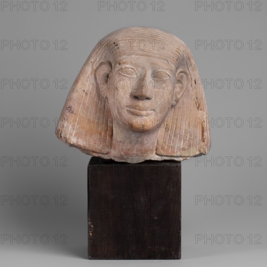 Head of a Man - Forgery, Unknown Role Classification of Creator, Author. Male portrait head. The face is slightly elongated with straight eyebrows and slightly slanting eyes. The upper eyelids are marked with rollers. The nose is wide, slightly angled, and upturned, with marked lobes and holes. The mouth is slightly protruding. The corners of the mouth are slightly turned up, giving the impression of an artificial smile. The chin is strongly marked. The ears are large, protruding, with elaborate earcups. A claft is worn low on the forehead, tucked behind the ears, and flows down to the shoulders. The folds of the claft are marked with parallel engravings. Traces of brown pigment in the folds. 1st half of the 20th century, Egypt. Sculpture, height 16.5 cm., A carved stone head mounted on a dark wooden block, showing a stylized face with a rounded forehead, prominent ears, and a subtle smile; the head has a striped, bobbed headdress with carved grooves and an overall worn, slightly rough...