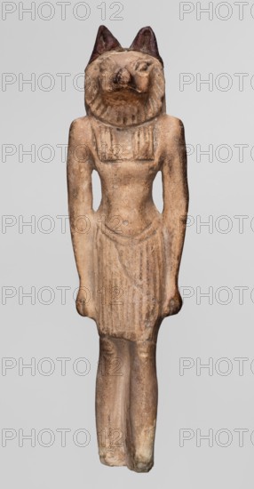 Amulet — Anubis. Anubis is depicted as a striding man with the head of a dog Canidae. Arms hang at his sides, hands clenched. Torso broad at the shoulders, narrowed at the waist. Pleated skirt with a loose belt, slightly lowered in the middle, reaches above the knees. Tripartite wig covers the head. A sort of mane is visible around the narrow, carefully modeled muzzle. Small, clumsily made ears are second-hand additions. Rear pillar reaches 2/3 of the figurine's height, with a drilled hole in its upper part. ca. 1069–525 BC; Third Intermediate Period–26th Dynasty. Origin: Egypt. Amulet. Height: 7.4 cm., A standing carved figure with an animal-like head and upright ears, depicting a human-shaped body wearing a short pleated skirt and a fitted upper garment, all in a weathered beige to light brown stone with darker brown accents on the ears and some shadowed areas; the surface shows rough texture and areas of lighter cream wear, with subtle variations of tan and ochre across the torso, a...