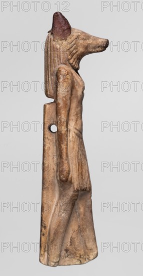 Amulet — Anubis. Anubis is depicted as a striding man with the head of a dog Canidae. Arms hang at his sides, hands clenched. Torso broad at the shoulders and narrowed at the waist. He wears a pleated skirt with a loose belt, slightly lowered in the middle, reaching above the knees. A tripartite wig covers the head. A sort of mane is visible around the narrow, carefully modeled muzzle. Small, clumsily made ears are second-hand additions. The rear pillar reaches 2/3 of the figurine's height, with a drilled hole in its upper part. Amulet, ca. 1069–525 BC Third Intermediate Period–26th Dynasty. Egypt. Height 7.4 cm., A carved figurine of a standing anthropomorphic animal with a long snout and upright pointed ears, shown in profile facing right, with a ribbed headdress or mane that falls down the back; the figure has a slender torso and arms held close to the body, a pleated skirt, and a flat back support with a circular hole. The surface shows weathered and worn textures with a palette of...