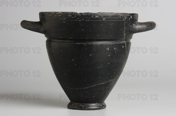 Skyphos, 1st quarter of the 6th century BC, Etruria; vessel; height 9.3 cm, width 9.5 cm., A matte black ceramic vessel with a flared rim and a rounded body narrowing to a small foot, showing a group of surface scuffs and small chips and a visible seam or repair line running vertically, with a few lighter speckles and worn areas; the vessel has a pair of short horizontal handles extending from opposite sides near the rim and sits against a light gray background, with visible shades of black, dark gray, light gray, and small hints of white where the surface is worn.