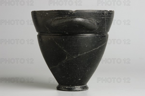 Skyphos, 1st quarter of the 6th century BC, Etruria, vessel, height 9.3 cm, width 9.5 cm, A dark, smooth vessel with a rounded rim and a tapered body sits centered against a light neutral background; the vessel is primarily black with subtle lighter scratches and small pale spots, showing a glossy surface and a couple of visible seams or hairline cracks, a short rounded foot at the base, and a shallow raised band near the top with a curved handle-like protrusion on one side, with overall tones of black, gray, and faint off-white on the background.