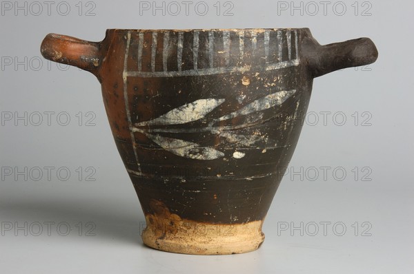 Black-finished skyphos with painted decoration on the rim, white decoration: A = B - laurel branch, IV waC, Etruria, vessel, height 9.7 cm, width 9.2 cm, A ceramic vessel with a tapered body and two short horizontal handles, shown against a pale gray background, featuring a dark brown to black surface with worn lighter areas and painted pale leaf-like shapes on its central body, a band of vertical pale stripes near the rim, and a lighter unglazed beige base that shows signs of wear and discoloration; the handles and upper rim show patches of reddish-brown and flaking, and the overall surface has spots where the dark finish has chipped to reveal lighter underlying material.