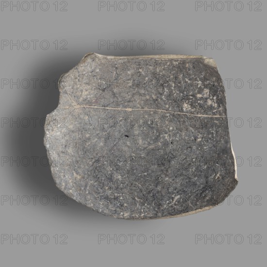 Fragment of a bucchero nero bowl with a flat bowl and a ring-shaped stem. On the inside of the bowl is scratched the Etruscan letter kh. On the bottom is engraved the complete Etruscan alphabet. Bucchero nero, ancient Etruscan vessel, 6th–5th century BC Archaic period. Origin: Etruria. Vessel fragment. Height 2.2 cm, width 10.5 cm., A roughly oval stone fragment with an uneven surface and jagged edges sits against a flat background. The stone shows various shades of gray and dark gray with lighter gray veins and small pale flecks dispersed across its face. A thin, lighter-colored line runs across the stone, and the edges reveal a beige to off-white tone. The background is a uniform medium gray with a soft darker gray shadow cast from the stone.