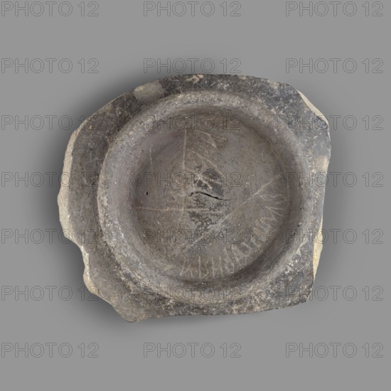 Fragment of a bucchero nero bowl. Bucchero nero; ancient Etruscan vessel, 6th–5th century BC Archaic period. Fragment with a flat bowl and a ring-shaped stem. On the inside of the bowl is scratched the Etruscan letter kh. On the bottom is engraved the complete Etruscan alphabet. Origin: Etruria. Vessel fragment. Height 2.2 cm; width 10.5 cm., A roughly circular stone object with an irregular outer edge rests on a plain gray background; the stone shows a recessed central basin with a raised rim and worn, rough surfaces, and the interior of the basin has visible scratches and incised lines forming a pattern; the stone displays a palette of gray tones with areas of darker charcoal gray, lighter gray, and patches of beige and brown on the fractured outer edges.