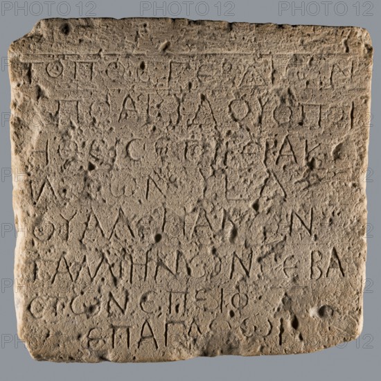 Greek dedicatory inscription, unknown author, Quadrangular limestone tablet with eight lines of Greek text. 25.06.257, Abydos, Greek inscription. Height 36.5 cm, width 39.0 cm, depth 7.1 cm., A roughly rectangular stone slab with a weathered, pitted surface and carved incised marks arranged in horizontal rows across the face, showing irregular edges and rounded corners; the stone displays a sandy beige to light brown color with subtle darker spots and shadows, and it is set against a plain gray background.