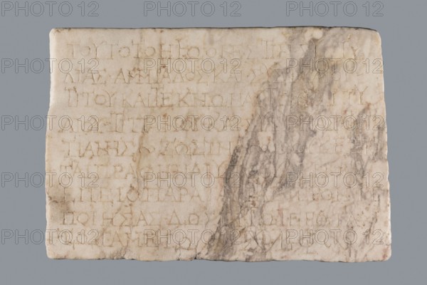 Titulus of the tomb of the Systilians, tombstone inscription in Greek, 2nd–3rd century, Phrygia; tombstone, Greek inscription; height 26.0 cm, width 38.0 cm, depth 4.5 cm, A rectangular stone slab with rounded corners showing surface carving and shallow grooves; the slab is primarily off-white and beige with areas of pale cream and warm tan, and a vertical streak of gray and darker gray marbling toward one side; weathered edges and small chips are visible along the perimeter, and faint carved lines form a grid-like pattern across the face; the surface shows patches of light brown discoloration and subtle shadowing in the carved depressions.
