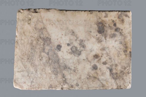 Titulus of the tomb of the Systilians, tombstone inscription in Greek, 2nd–3rd century, Phrygia; material: tombstone; dimensions: height 26.0 cm, width 38.0 cm, depth 4.5 cm., A rectangular stone slab with a rough, worn surface showing a mix of off-white, beige, light tan, gray, and darker gray spots and smudges, with irregular edges and small chips, several rounded darker stains, a few faint veins and streaks across the surface, and an even gray background behind the slab.