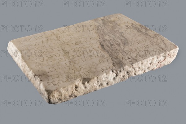 Titulus of the tomb of the Systilians, tombstone inscription in Greek, 2nd–3rd century, Phrygia; tombstone, height 26.0 cm, width 38.0 cm, depth 4.5 cm., A rectangular stone slab rests on a plain background, showing a worn, uneven top surface and rough, eroded edges. The top has shallow carved markings arranged in rows and an irregular darker stain or discoloration across part of it. The stone's colors include shades of beige, off-white, light gray, and areas of darker gray and brownish tones, with some subtle reddish-brown patches along the sides. The texture ranges from smoother, slightly polished areas on the top to coarse, pitted, and crumbly surfaces along the edges.