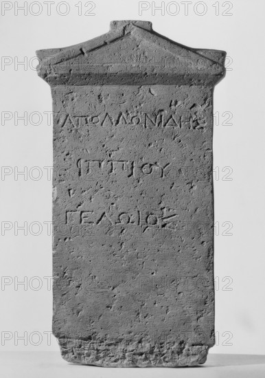 Tombstone of Apollonides with Greek inscription. Rectangular slab with three lines of Greek inscription, topped with a triangular tympanum. Tombstone of Apollonides, son of Hippias of Gela, 300–250 BC; Ptolemaic period. Found at Zawiyet el-Mayetin near Minia, Kom el Ahmar. Greek inscription. Height 58.5 cm, width 32 cm, depth 9 cm, weight 25.5 kg., A single upright stone slab with a triangular pediment at the top, showing a rough, pitted surface and weathered edges; the slab appears textured with small pits and shallow grooves, and there are carved linear depressions across its face; the base is irregular and worn, with a chipped lower edge and a rough, uneven bottom; the visible colors are shades of gray and off-white, with subtle darker gray shadows accentuating the surface texture.