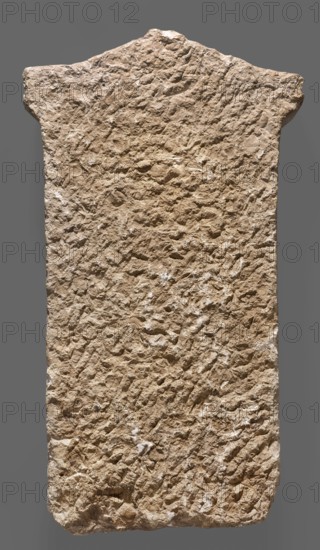 Tombstone of Apollonides with Greek inscription. Rectangular slab with three lines of Greek inscription, topped with a triangular tympanum. Tombstone of Apollonides, son of Hippias of Gela, 300–250 BC; Ptolemaic period. Found at Zawiyet el-Mayetin near Minia, Kom el Ahmar. Greek inscription. Height 58.5 cm, width 32 cm, depth 9 cm, weight 25.5 kg., A tall, roughly rectangular stone slab with an uneven top edge and irregular sides, showing a heavily textured, pitted surface with a rough, granular appearance; the slab is beige to light brown with small areas of lighter off-white patches and subtle darker brown shadows, and it is set against a flat gray background.