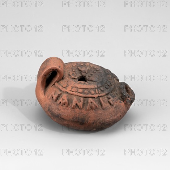 Lamp with the Greek inscription C???? ??? ????? May he be my saviour; crosses at the beginning and end of the inscription. On the disc four Greek letters alpha arranged in the sign of the cross. Unknown Aswan workshop; 5th/6th century AD dating revised to 6th–7th century. The lamp's container is partly wheel-turned. The burner is very short and small. A small, band-type vise. The arms around the olive hole feature a beaded ornament enclosed in two circles. Below it is a convex imprinted inscription: Jesus, my savior. At its beginning and end are equal-armed crosses. The disk bears four convex letters Alpha arranged in the shape of a cross. The lamp's front surface is charred, and the underside is stained with oil. Found in Edfu; oil lamp; height 4.4 cm, width 7.6 cm, depth 6.8 cm., A small rounded clay object in shades of reddish-brown and dark brown rests on a light gray surface against a pale gray background; it has a curled handle-like protrusion on one side and a central circular d...