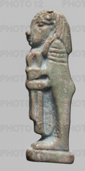 Amulet — Imset. Figurine of Imset one of the Four Sons of Horus, worked in relief; shown in left profile as a mummy-like figure with a human head covered by a wig, holding a folded cloth in front of him. Smooth back with two holes for attachment. Part of a set depicting the Four Sons of Horus. Late Period–Ptolemaic Period, 664–30 BC, Egypt. Amulet; height 4.6 cm, width 1.5 cm., A small statuette of a standing humanlike figure shown in profile with a rounded head and stylized facial features, short hair or headdress, arms folded across the chest holding an object, a long garment draped to the feet, a rectangular base beneath the feet, and a small hole at the top of the head; the surface shows a weathered, slightly pitted texture with a patina of bluish-green, green, brown, tan, and gray tones.