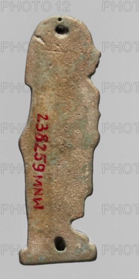 Amulet — Imset; Figurine of Imset one of the four Sons of Horus, worked in relief; shown in left profile with a mummy-like body and a human head covered with a wig; holds a scroll of fabric in front; back is smooth with two holes for attachment. Part of a set depicting the four Sons of Horus. Date: 664–30 BC, Late Period–Ptolemaic Period, Egypt. Amulet; height 4.6 cm, width 1.5 cm., A flat, vertically oriented object shaped like a stylized standing figure with a rounded head, angular shoulders, and undulating sides, shown against a plain light gray background; the surface is rough and pitted with uneven texture and areas of discoloration; visible colors include tan, brown, greenish-blue patina, and small darker spots; there is a circular hole near the top and another near the bottom; the outline includes a small protruding nose-like feature and a stepped base.