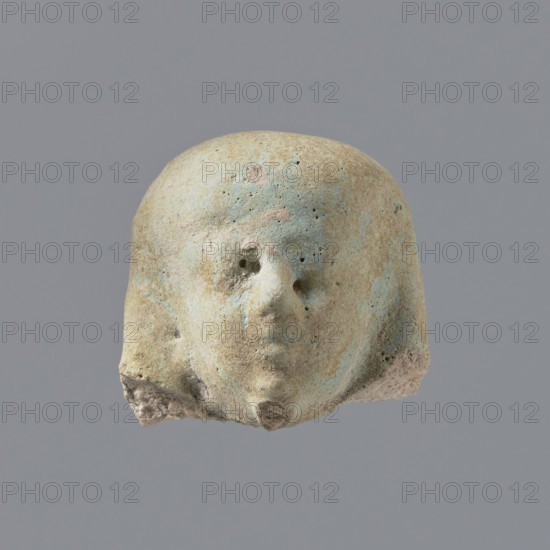 Fragment of a shabti. Fragmentary head of a shabti figurine with indistinct facial features, in a smooth wig. Celadon glaze. Ptolemaic Period, 332–30 BC. Found at El-Kantara. Shabti; height 2.3 cm., A worn pale beige object with patches of light blue and hints of pink that resembles a rounded head with a few small holes where facial features are, a subtle protruding nose shape and an indistinct mouth area, a few rough textured spots and small pits across the surface, and a darker beige irregular base at the bottom, set against a uniform gray background.