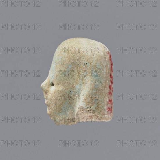 Fragment of a shabti;, Head of a shabti figurine with indistinct facial features, in a smooth wig. Celadon glaze. Ptolemaic Period, 332–30 BC. Found at El-Kantara. Shabti; height 2.3 cm., A worn small sculpted head shown in profile against a plain gray background, with a rounded top and a simplified facial outline including a protruding nose and a small recessed eye; the surface appears rough and pitted with areas of pale beige and light blue-green patina, and a vertical strip of reddish pigment along the rear edge.