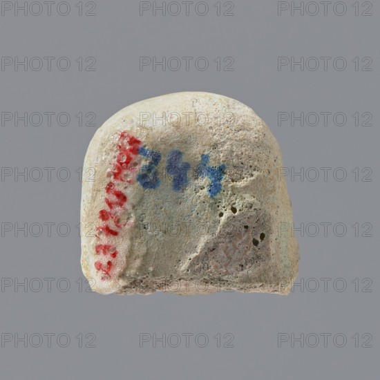 Fragment of a shabti, Head of a shabti figurine with indistinct facial features, in a smooth wig. Celadon glaze. 332–30 BC; Ptolemaic Period. Found at El-Kantara. Shabti; height 2.3 cm., A small rounded stone or pebble with a rough, porous surface shown against a plain gray background. The stone is primarily light beige and off-white with areas of pale gray and faint greenish tints. There is a vertical streak of red pigment along one side and a few splotches of blue pigment near the top center. The surface shows pits, small holes, and irregular textures with a slightly worn bottom edge.