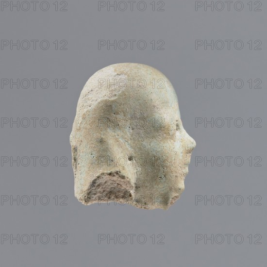Fragment of a shabti, heads, grave goods, wigs. Head of a shabti figurine with indistinct facial features, in a smooth wig. Celadon glaze. 332–30 BC; Ptolemaic Period. El-Kantara, shabti, height 2.3 cm., A worn sculpted head viewed in profile against a plain gray background, showing a rounded top, a defined nose and forehead, and a rough, eroded lower edge; the surface has a weathered texture with patches of pale beige, light tan, muted green, and hints of bluish-gray, and there are small pits and irregularities across the form.