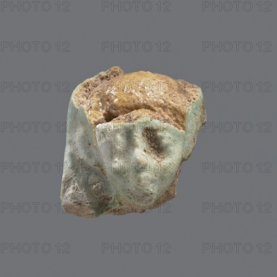 Fragment of a shabti, head of a shabti figurine with carefully rendered facial features in a smooth wig, celadon glaze. Ptolemaic Period, 332–30 BC; found at El-Kantara. Shabti, height 2.4 cm., A small weathered fragment resembling a stylized face sits against a plain gray background, with a worn surface showing a mix of pale turquoise, light blue, beige, tan, and brown tones; the fragment has rounded, smoothed contours with indentations where eye and mouth shapes appear, and a rough, granular texture with some glossy, slightly reflective patches and areas of encrusted debris.