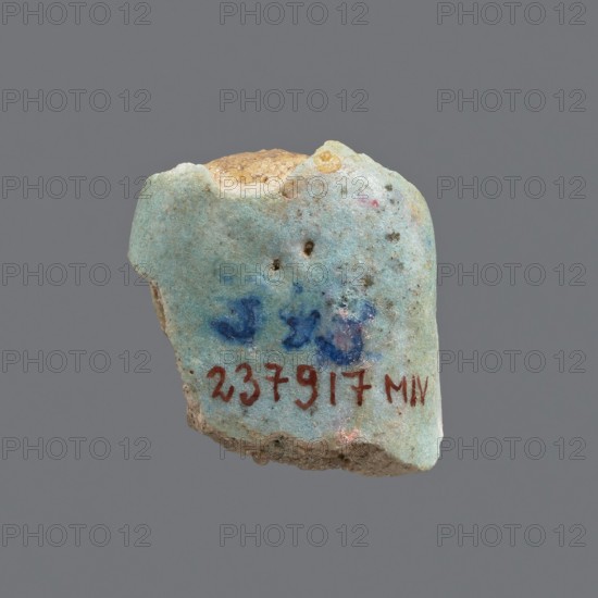 Fragment of a shabti; head of a shabti figurine with carefully rendered facial features in a smooth wig. Celadon glaze. Ptolemaic Period, 332–30 BC. Found at El-Kantara. Shabti; height 2.4 cm., A small irregular fragment of a hard material with a worn, rough surface in shades of pale turquoise and light blue, with areas of darker blue spots and a few tiny dark brown specks; the top edge shows exposed beige and tan inner material, and the lower edge reveals a rough, sandy brown interior; there are traces of faint pinkish spots and a small patch of yellow near the top, set against a plain gray background.