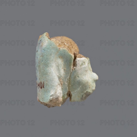 Fragment of a shabti. Head of a shabti figurine with carefully rendered facial features, in a smooth wig. Celadon glaze. Ptolemaic Period, 332–30 BC. Found at El-Kantara. Shabti; height 2.4 cm., A small irregular object with a rough textured surface, showing a combination of pale turquoise, light blue, beige, tan, and off-white colors, with areas that appear worn and slightly glossy; the object has rounded and jagged contours, a bulbous tan section partially enclosed by flaking turquoise-blue layers, and a few small pits and cracks visible across its surface.