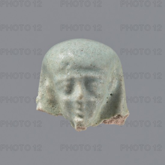 Fragment of a shabti. Head of a shabti figurine with carefully rendered facial features, in a smooth wig, with a thin beard. Celadon glaze. Ptolemaic Period, 332–30 BC. El-Kantara place of discovery. Shabti; height 2.5 cm., A small sculpted head fragment with a rounded top and stylized facial features, showing a forehead, recessed eyes, a subtle nose, and a suggestion of a mouth and chin; the surface is worn and pitted with rough broken edges at the lower sides and bottom; the piece is predominantly a pale bluish-green with speckles of darker green and tiny brown spots, and there are areas of exposed beige or light tan material where the surface is chipped; the background is a uniform medium gray.