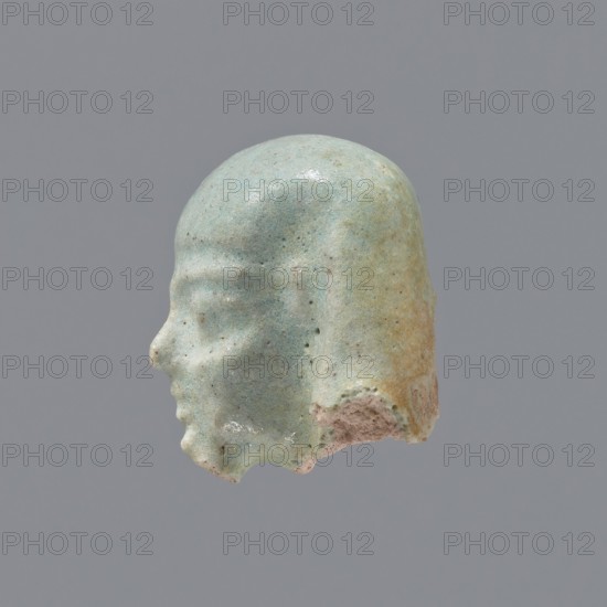 Fragment of a shabti; head of a shabti figurine with carefully rendered facial features, in a smooth wig, with a thin beard. Celadon glaze. Ptolemaic Period, 332–30 BC; El-Kantara. Shabti, height 2.5 cm., A small sculpted head shown in profile with a smooth rounded top and a defined forehead, brow ridge, nose, lips, chin, and a stylized hairstyle that falls down the back of the head, all rendered in a weathered pale green with areas of light blue and touches of beige and tan where the surface is worn, set against a plain medium-gray background.