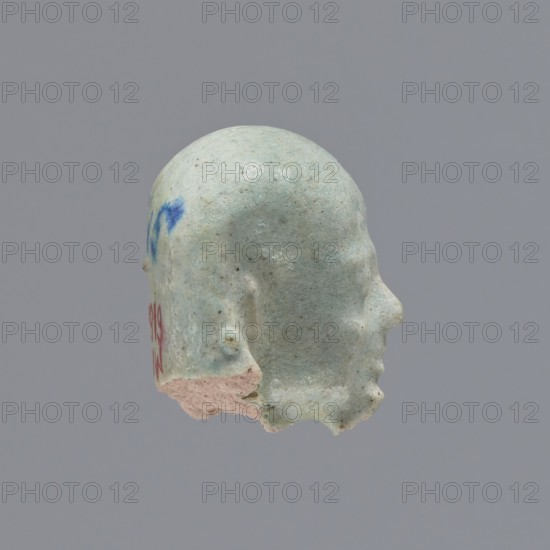 Fragment of a shabti. Head of a shabti figurine with carefully rendered facial features, in a smooth wig, with a thin beard. Celadon glaze. 332–30 BC; Ptolemaic Period. El-Kantara. Shabti, height 2.5 cm., A profile view of a small sculpted head fragment with a rounded top and defined nose, lip, and chin contours, showing a textured surface with chips and missing areas; the object is primarily pale green with areas of off-white and a few patches of blue and pink, set against a uniform gray background.
