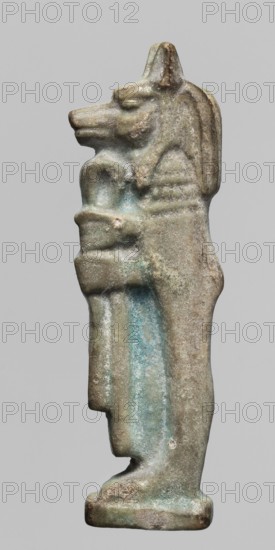 Amulet - Duamutef. Relief figurine of Duamutef, one of the Sons of Horus. A mummy-like figure with a dog's head covered with a wig, holding a scroll of fabric in front of himself. The back is smooth with two holes for attachment. Part of a set of four Sons of Horus. Late Period–Ptolemaic Period, 664–30 BC, Egypt. Amulet; height 4.6 cm, width 1.4 cm., A small weathered figurine of an animal-headed standing figure shown in profile against a plain light gray background, with a pronounced snout and pointed ear, a mane-like ridge along the back of the head, and arms folded across the chest holding a long vertical object; the surface is rough and pitted with worn edges, showing a patina of greenish-blue, pale turquoise, gray, beige, and areas of brownish tan.
