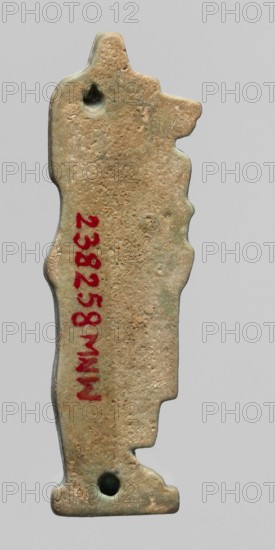 Amulet - Duamutef. Relief figurine of Duamutef, one of the Sons of Horus. A mummy-like figure with a dog's head covered with a wig, holding a folded linen scarab? or roll of fabric in front of himself. The back is smooth with two holes for attachment. Part of a set of four Sons of Horus. Material: amulet; date: 664–30 BC, Late Period–Ptolemaic Period, Egypt. Dimensions: height 4.6 cm, width 1.4 cm., A vertically oriented worn object with an irregular silhouette resembling a standing figure, showing a rough textured surface in shades of beige, tan, and light brown with areas of greenish patina; a few small dark green spots and darker indentations are visible near the top and bottom, and a group of red markings appears along the object's midsection; the background is a uniform light gray.