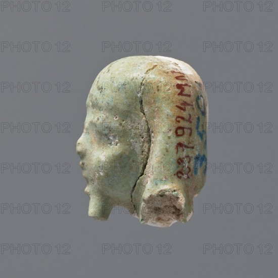 Fragment of a shabti. Head of a shabti figurine with rather carelessly rendered facial features, in a smooth wig and a thin beard. Celadon glaze. 332–30 BC; Ptolemaic Period. El-Kantara. Shabti, height 2.6 cm., A small sculpted head shown in profile against a neutral gray background, with a worn surface of pale green and turquoise tones mixed with areas of light beige and off-white, and traces of darker green and brown discoloration; the facial features are rounded and simplified with a prominent nose and small lips, a chipped lower edge, and a visible hair or headdress form that follows the skull and ends near the base; the texture appears grainy and eroded with cracks and small pitted areas, and there are a few spots of reddish and blue pigment on the side.