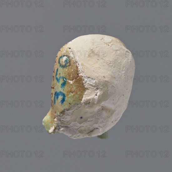 Fragment of a shabti. Head of a shabti figurine with rather carelessly rendered facial features, in a smooth wig and a thin beard. Celadon glaze. 332–30 BC; Ptolemaic Period. Found at El-Kantara. Shabti; height 2.6 cm., A roughly oval object with an uneven, textured surface, showing a contrast between a creamy off-white area and a weathered greenish-brown area; the greenish-brown section has traces of blue marks and small patches of red, while the off-white area appears chalky with rough, pitted texture and some small darker specks; lighting creates soft shadows and highlights that emphasize the surface irregularities, and the background is a uniform muted gray.