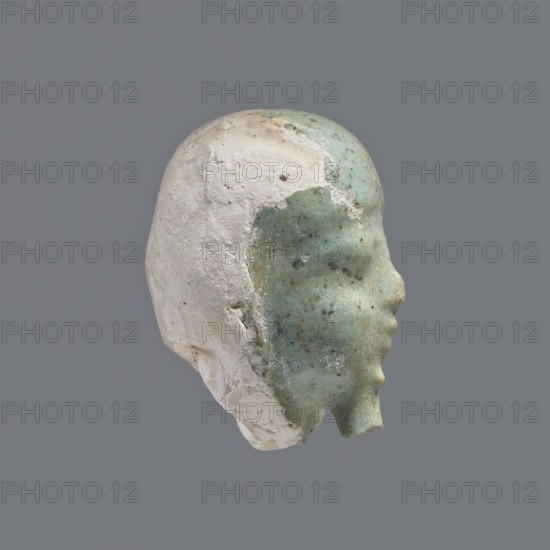 Fragment of a shabti. Head of a shabti figurine with rather carelessly rendered facial features, in a smooth wig and a thin beard. Celadon glaze. 332–30 BC; Ptolemaic Period. Found at El-Kantara. Shabti, height 2.6 cm. Provenance: grave goods; wigs., A sculpted head fragment shown in profile with a smooth face surface that is predominantly greenish-blue mottled with darker speckles and patches, while the rear and top of the head are a rough, chalky white with textured, flaking areas; the background is a uniform medium gray.