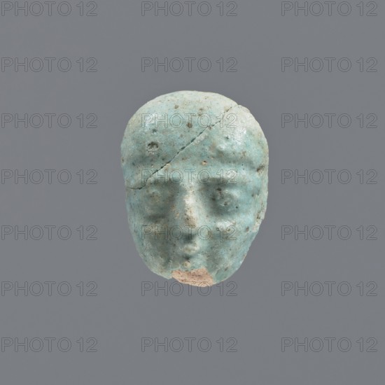 Fragment of a shabti, grave goods, wigs. Face of a shabti figurine with carefully rendered facial features, with the beginning of a smooth wig visible above the forehead. Bluish glaze. 332–30 BC Ptolemaic Period, El-Kantara, shabti, height 2.2 cm, A worn sculpted head with a rounded shape, showing a few surface cracks and chips, rendered mainly in pale turquoise with patches of light beige where material is missing, subtle darker speckles across the surface, faintly defined eye sockets, a suggestion of a nose and mouth, and an overall matte textured appearance against a solid gray background.