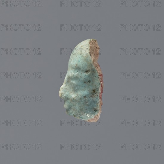 Fragment of a shabti, grave goods, wigs. Face of a shabti figurine with carefully rendered facial features, with the beginning of a smooth wig visible above the forehead. Bluish glaze. 332–30 BC; Ptolemaic Period. El-Kantara, shabti, height 2.2 cm., A small sculpted fragment of a human profile facing left, with a textured surface in shades of pale turquoise and light blue, a worn beige area along the back edge, and a thin strip of muted red visible on the rear side, set against a uniform gray background.