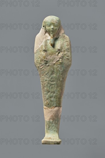Ushabti, Mummy-shaped figurine, standing on a base with the back resting on a pillar. Arms crossed on the chest with hoes in both hands; hands indistinctly marked. Wearing a long, smooth, three-part wig with ears uncovered. Small, round face with slightly blurred details and a long, straight beard adhering to the neck. Painted inscription on the front of the figurine's legs between engraved vertical lines text illegible. Mummy-shaped figurine used as grave goods; shabti. Ptolemaic period, 332–30 BC; findspot El-Kantara. Height 16.3 cm., A small elongated statue resembling a wrapped figure with a rounded head and a narrow rectangular base, showing a worn surface texture with areas of roughness and flaking; the form tapers from broader shoulders to a slimmer lower body and stands upright against a plain background. The visible colors include shades of green, pale green, beige, tan, and hints of gray, with some lighter patches and darker weathered areas. The head appears smoother and slig...