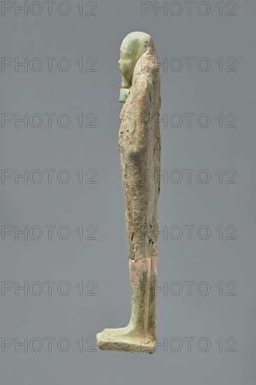 Ushabti, Mummy-shaped figurine standing on a base with the back resting on a pillar. Arms crossed on the chest, hands indistinctly marked, holding hoes in both hands. Long, smooth, three-part wig with ears uncovered. Small, round face with slightly blurred details and a long, straight beard adhering to the neck. Painted inscription on the front of the figurine's legs between engraved vertical lines text illegible. 332–30 BC, Ptolemaic period. Find location: El-Kantara. Shabti, height 16.3 cm., A slender vertical figure viewed in profile, with a rounded head and a simplified face, stands with arms close to the body and an elongated torso and legs; the surface shows a textured, weathered patina with areas of greenish and pale beige tones and hints of brown, and the base and lower legs display smoother, slightly lighter patches, all set against a plain gray background.