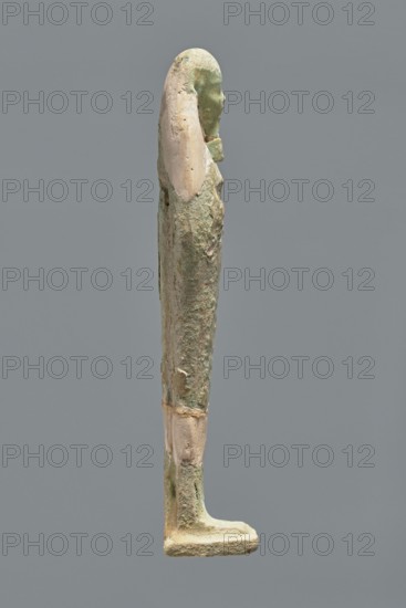 Ushabti, Mummy-shaped figurine, standing on a base with the back resting on a pillar. Arms crossed on the chest, hands indistinctly marked, holding hoes in both hands. Long, smooth three-part wig; ears uncovered. Small, round face with slightly blurred details; long, straight beard adhering to the neck. Painted inscription on the front of the figurine's legs between engraved vertical lines text illegible. Mummy-shaped figurine shabti, Ptolemaic period, 332–30 BC; find location El-Kantara. Height 16.3 cm., A slender standing statue in profile with a long headdress and defined facial features, showing a smooth head and elongated body with arms close to the sides, rendered in a weathered surface combining pale beige and green tones with rough and smoother patches, set against a plain medium gray background.